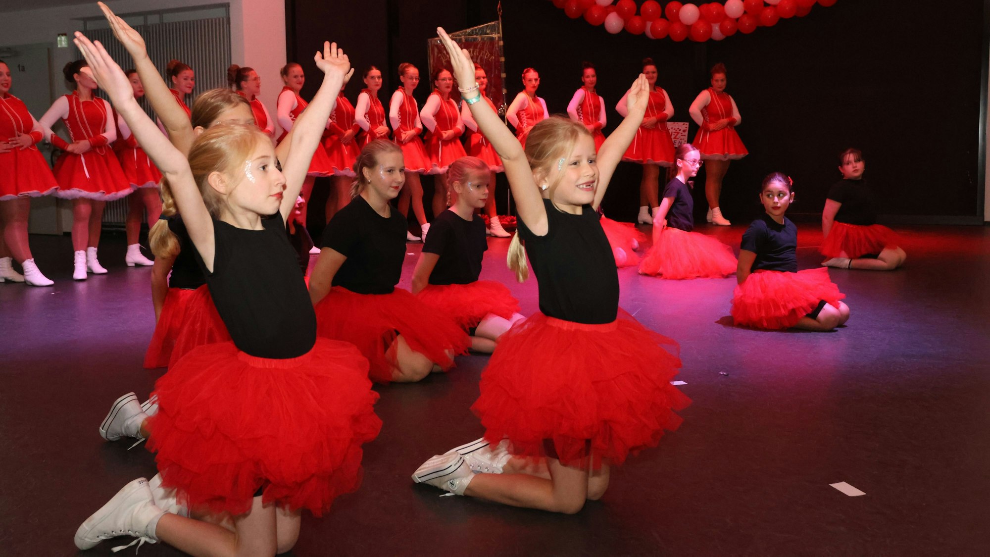 Im roten Tüll Rock und schwarzem Oberteil, knien und sitzen junge Mädchen auf der Bühne.