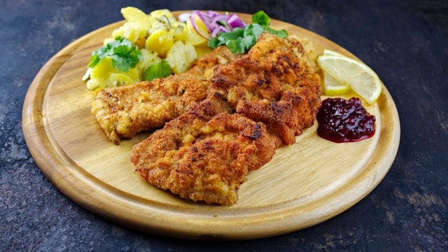 Knuspriges Schnitzel mit Kartoffelsalat, Preiselbeeren und Zitrone auf rustikalem Holzbrett.
