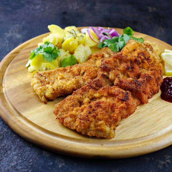 Knuspriges Schnitzel mit Kartoffelsalat, Preiselbeeren und Zitrone auf rustikalem Holzbrett.