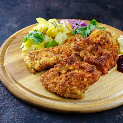 Knuspriges Schnitzel mit Kartoffelsalat, Preiselbeeren und Zitrone auf rustikalem Holzbrett.