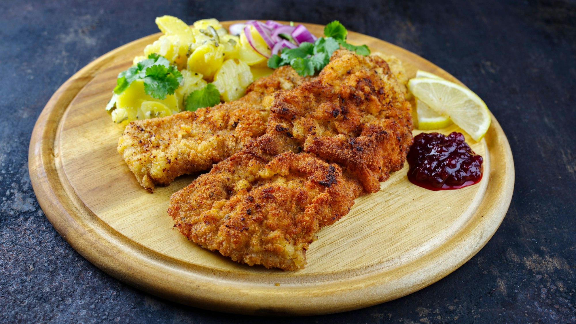 Knuspriges Schnitzel mit Kartoffelsalat, Preiselbeeren und Zitrone auf rustikalem Holzbrett.