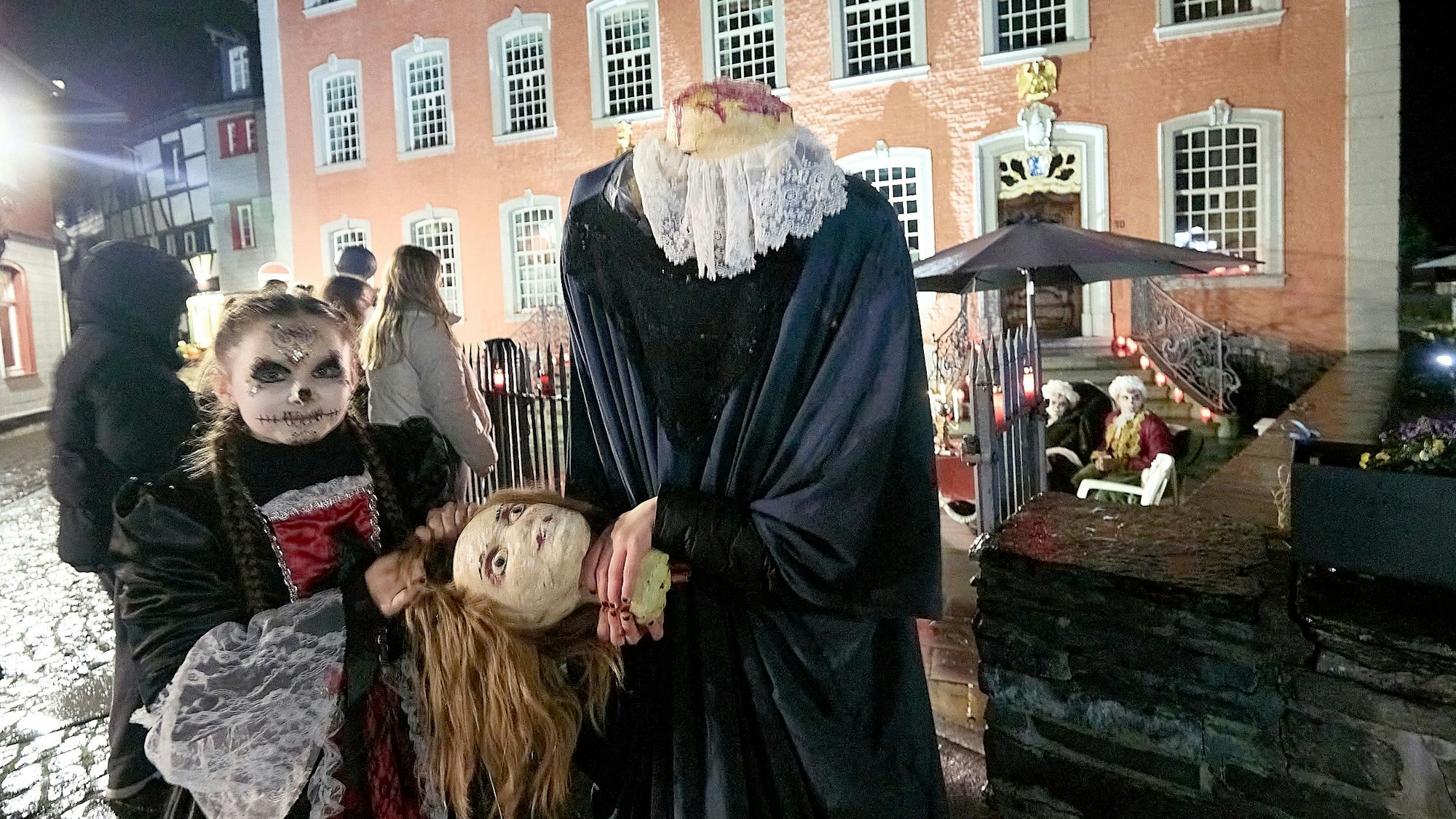 Zwei Figuren stehen vor dem Roten Haus in Monschau und halten einen Kopf in der Hand, der der einen Figur fehlt.