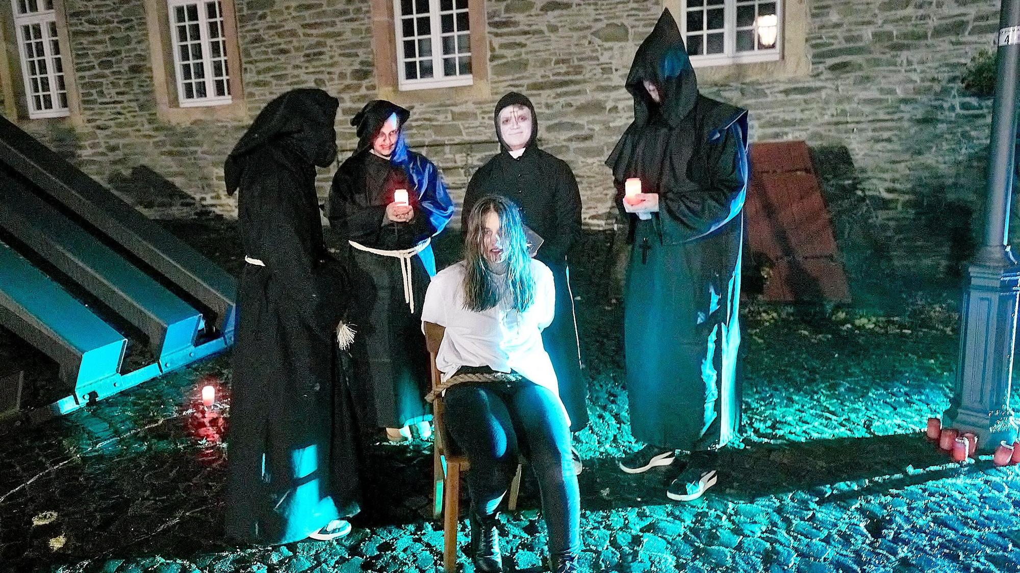 Mehrere Personen stehen in dunklen Roben um eine Frau herum, die auf einem Stuhl sitzt und festgebunden ist.