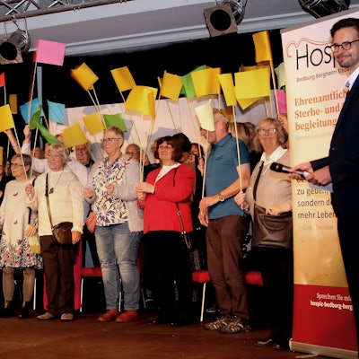 Das Bild zeigt mehrere Personen mit bunten Fähnchen auf einer Bühne.