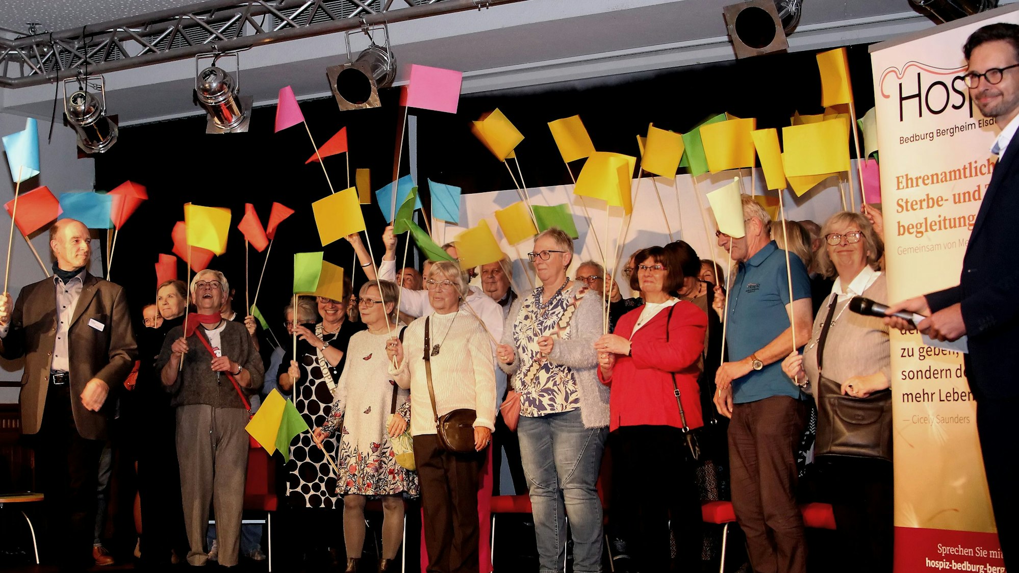 Das Bild zeigt mehrere Personen mit bunten Fähnchen auf einer Bühne.