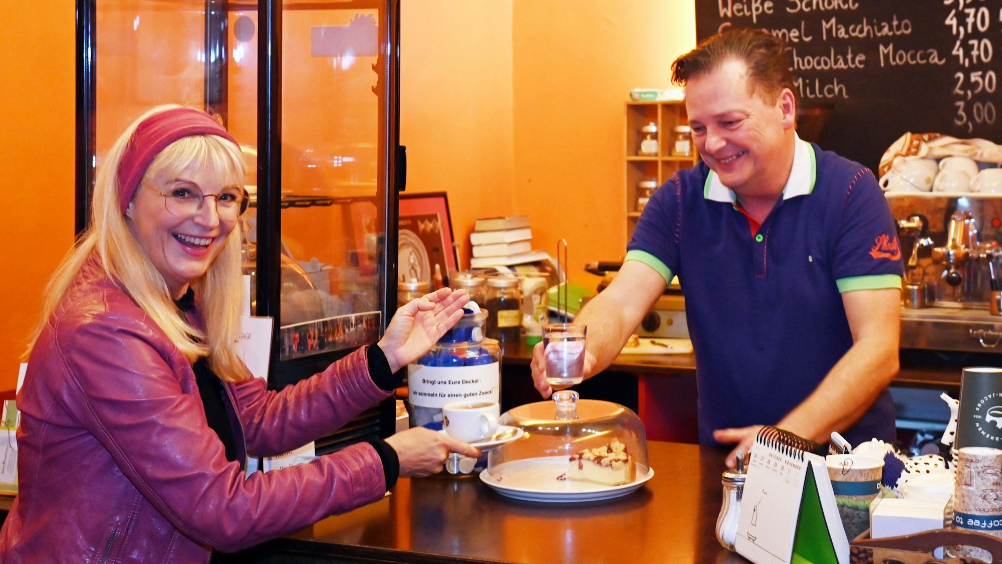 Eine blonde Frau mit rotem Haarband nimmt ihren Kaffee entgegen von einem Mann mit braunem kurzen Haar und blauem T-Shirt.