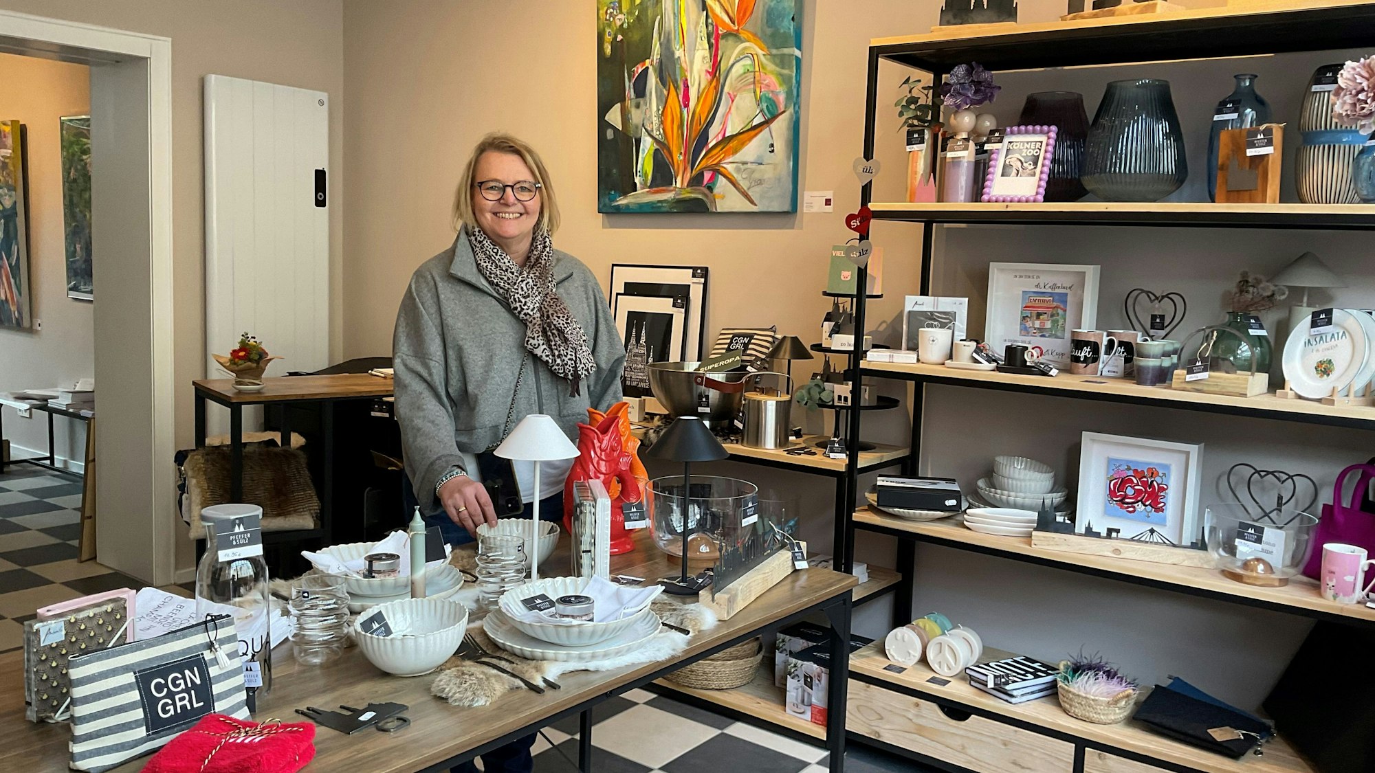 Eine Frau mit blondem Haar steht hinter einem Tisch in einem Ladenlokal, in dem allerlei Dekoartikel ausgestellt sind.