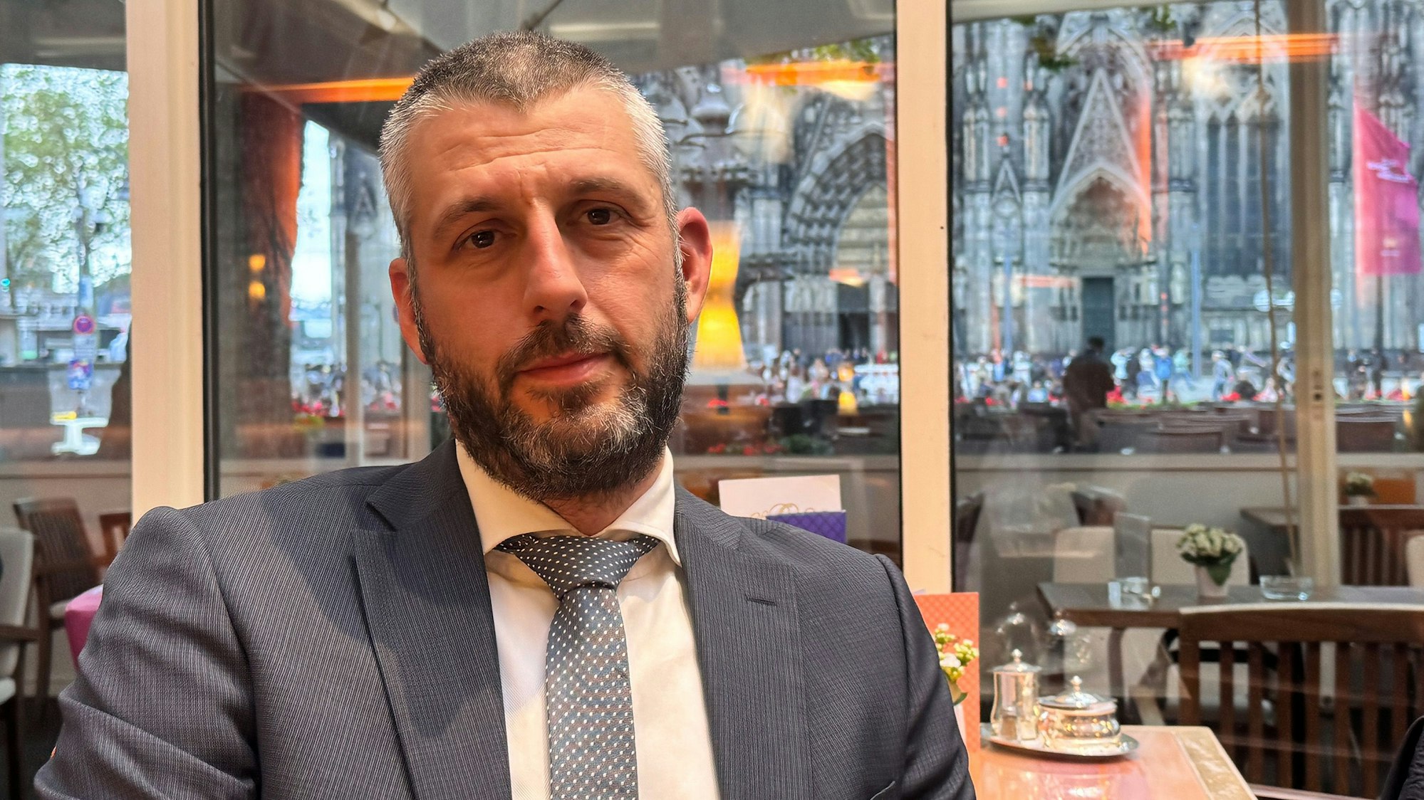 Auf dem Bild ist ein Mann mit Bart und in einem grauen Anzug in einem Kölner Café zu sehen.