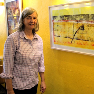 Eine Frau mit schulterlangem Haar steht neben einem Bild, das an einer gelben Wand hängt.