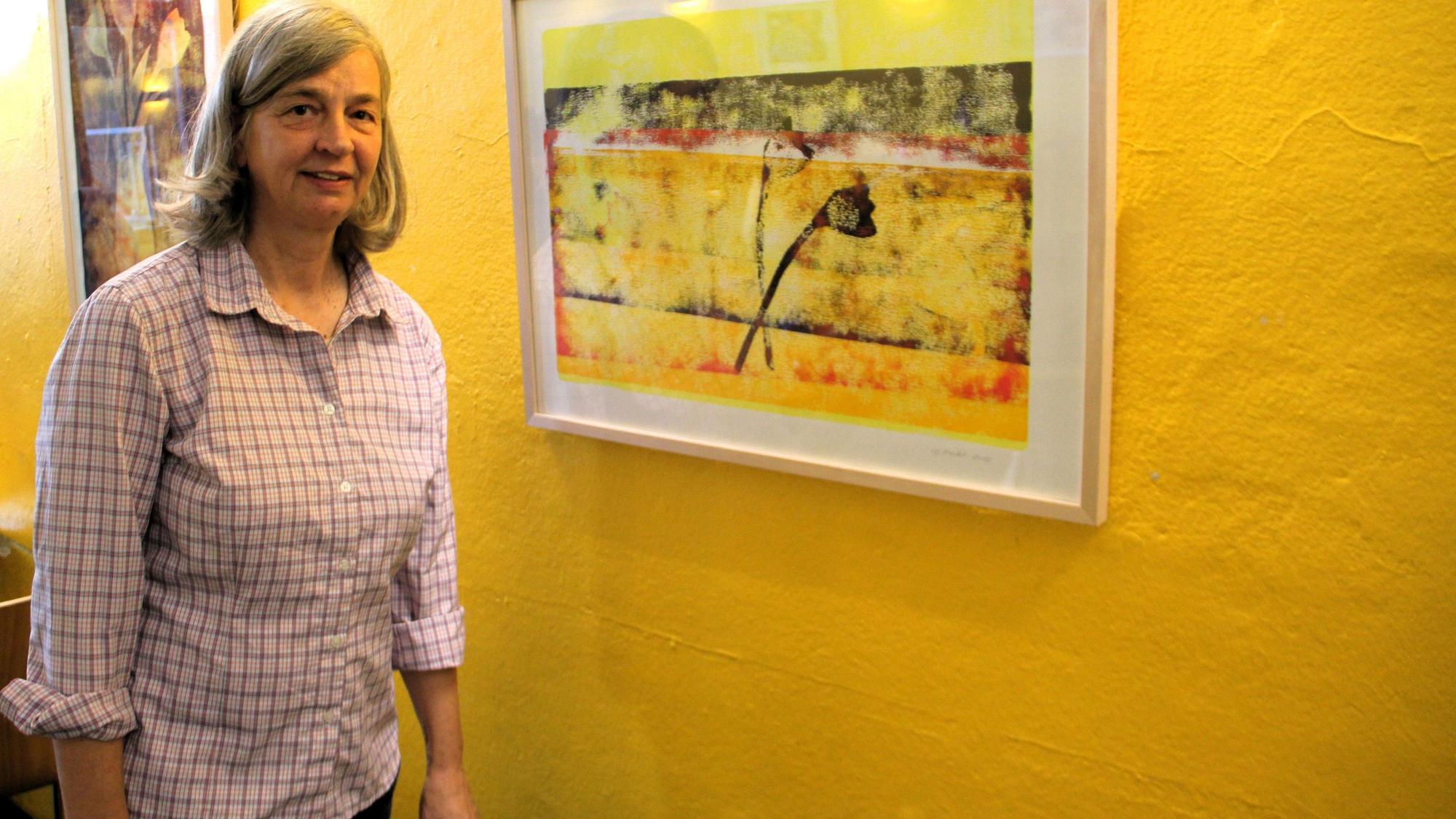 Eine Frau mit schulterlangem Haar steht neben einem Bild, das an einer gelben Wand hängt.