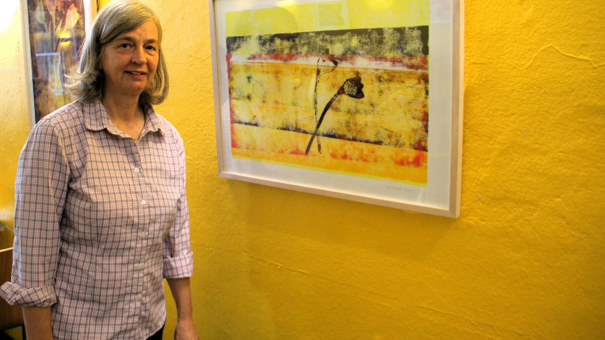 Eine Frau mit schulterlangem Haar steht neben einem Bild, das an einer gelben Wand hängt.
