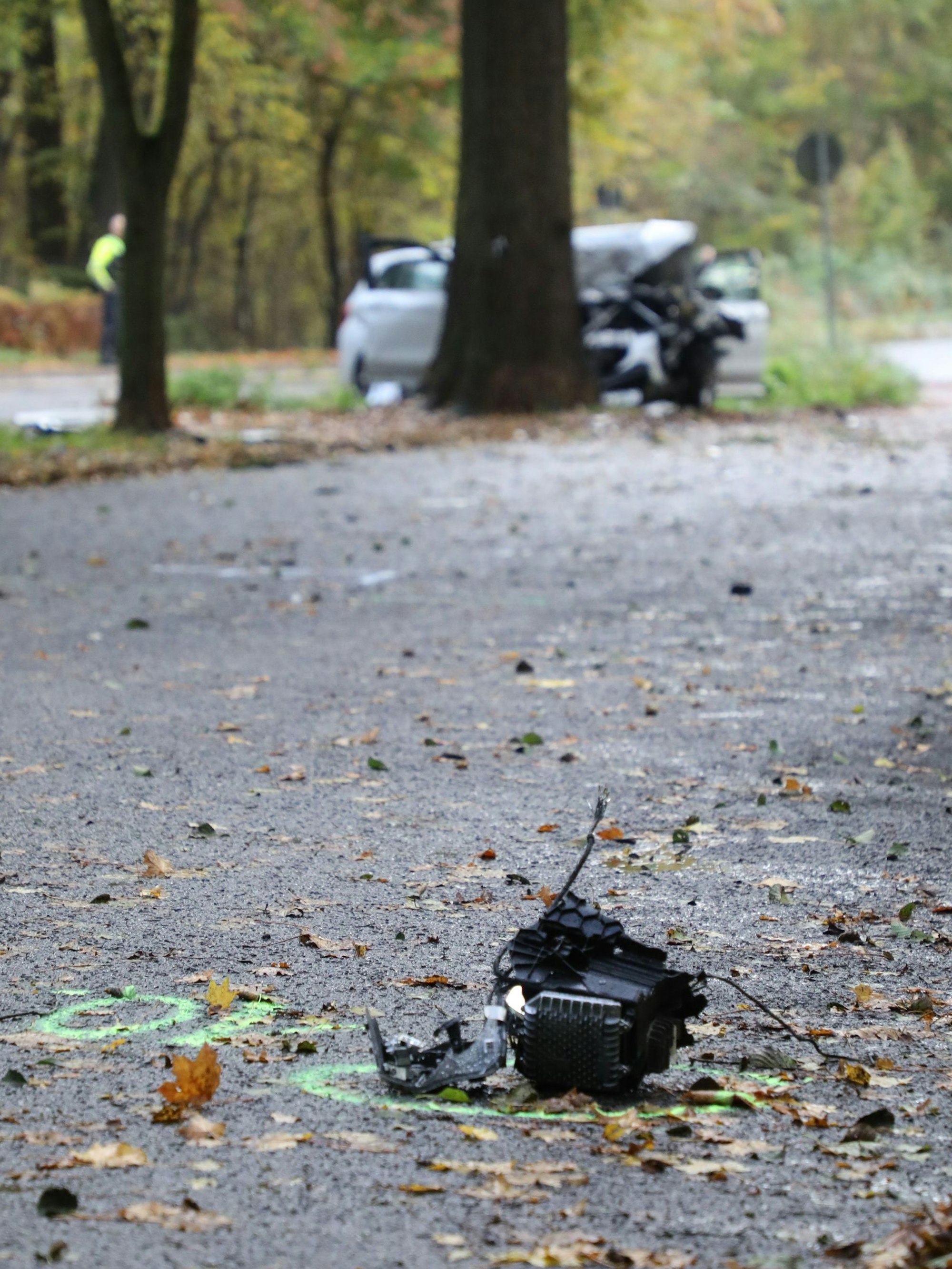 Ein Fahrzeugteil liegt auf dem Wanderparkplatz bei Rösrath-Kleineichen. Im Hintergrund ist ein Unfallwagen vor einem Baum zu sehen.