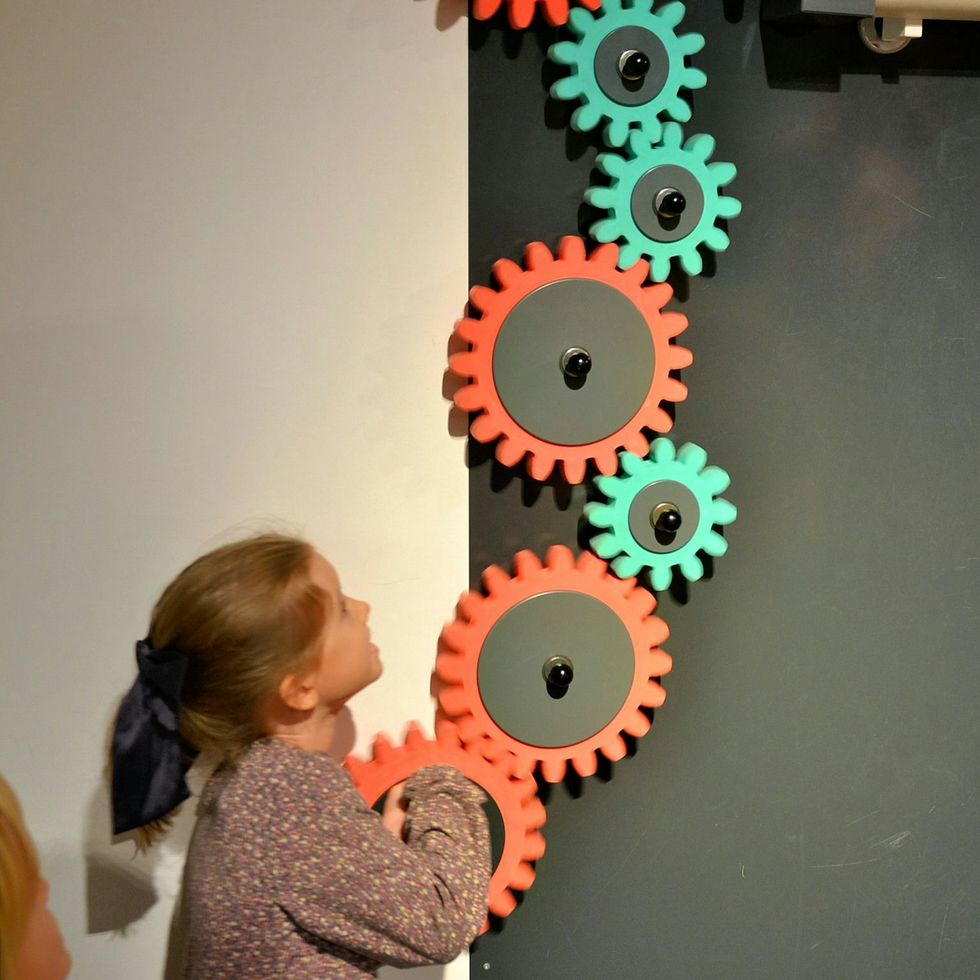 Ein Mädchen dreht an ineinander greifenden Zahnrädern, mit denen die Kugel transportiert wird.