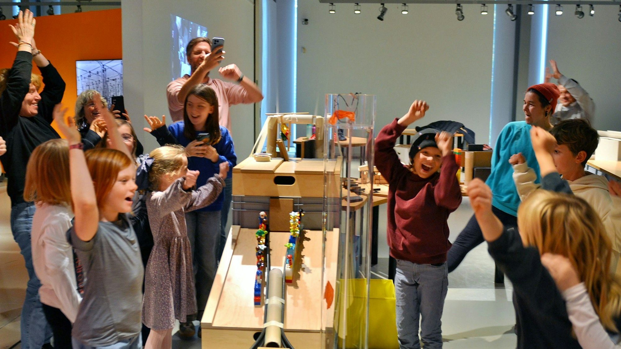 Kinder und Erwachsene stehen jubelnd um eine Kegelbahn herum. Manche haben die Arme nach oben gerissen.