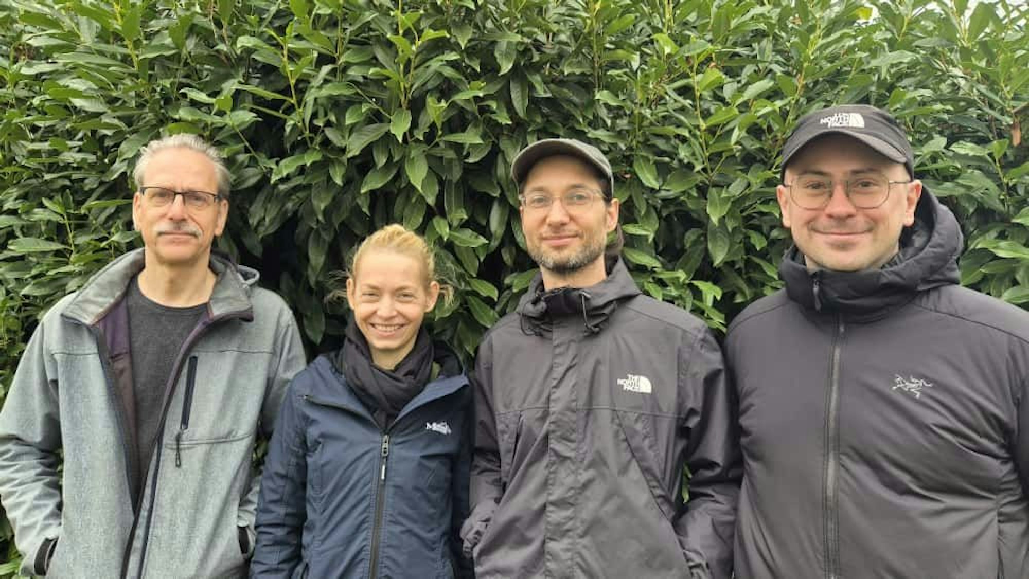 Drei Männer und eine Frau stehen nebeneinander vor einer grünen Hecke,