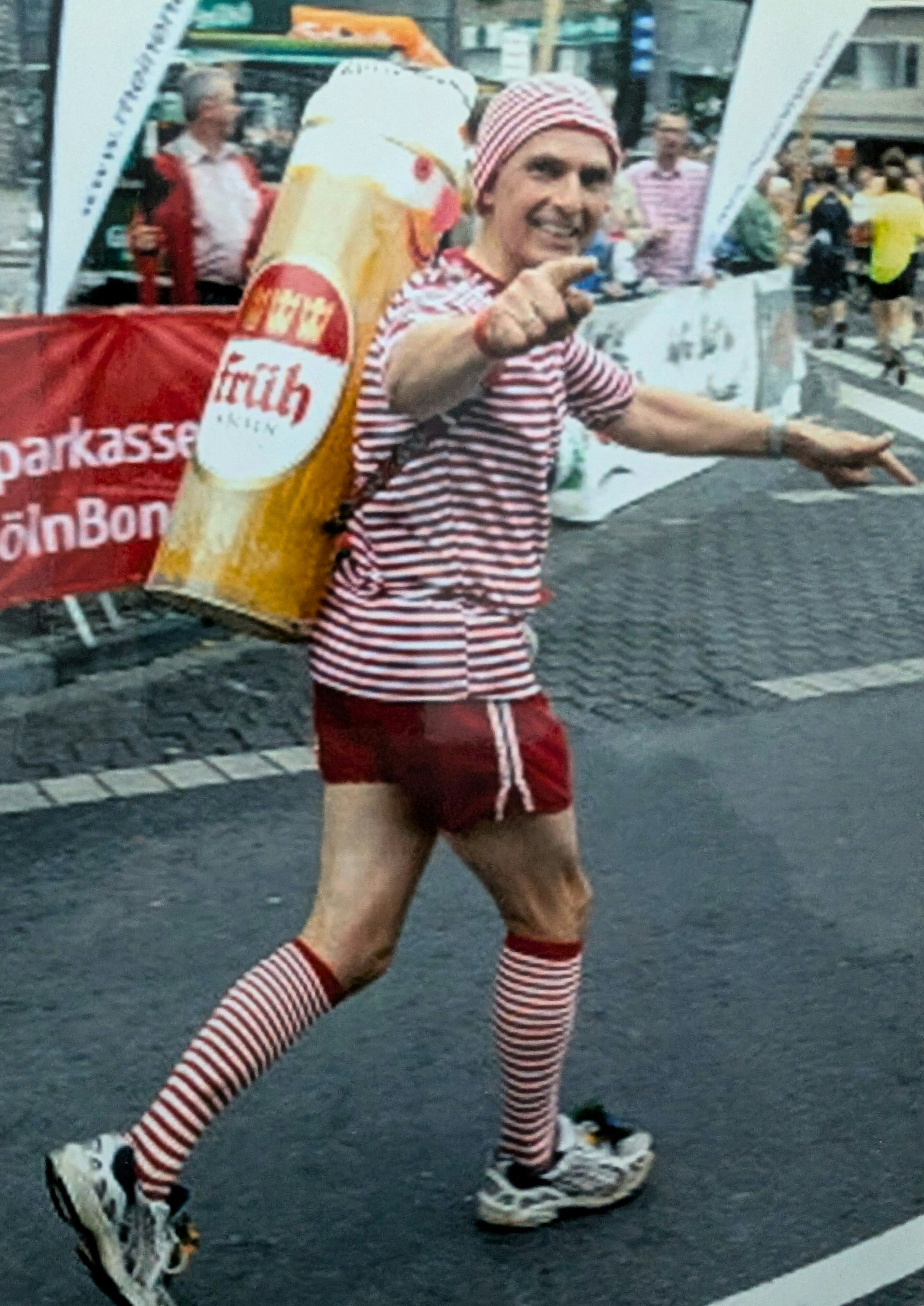 Ein Mann läuft in gestreiftem Trikot mit Kölschglas auf dem Rücken eine Straße entlang.