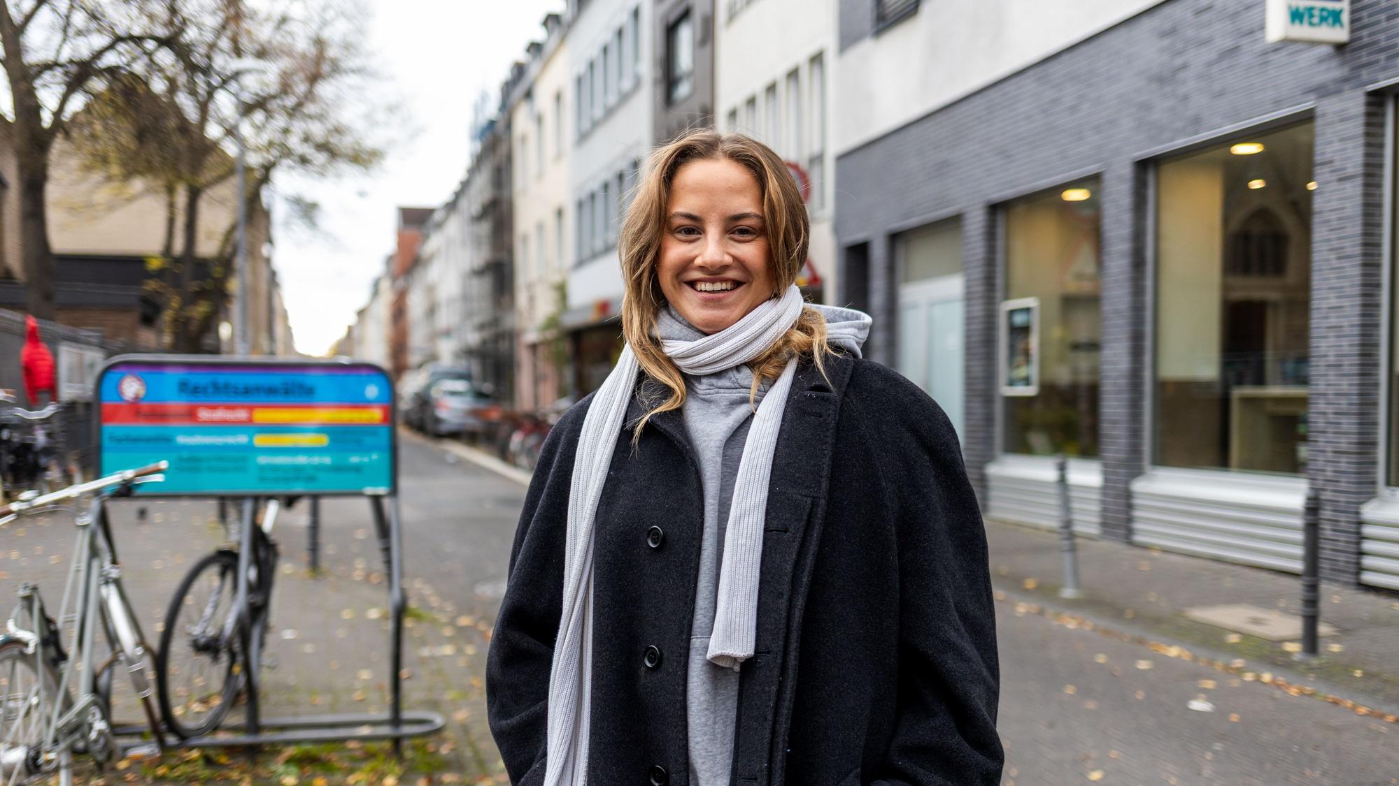 Eine junge Frau mit dickem schwarzen Mantel und weißem Schal steht auf dem Bürgersteig der Venloer Straße.