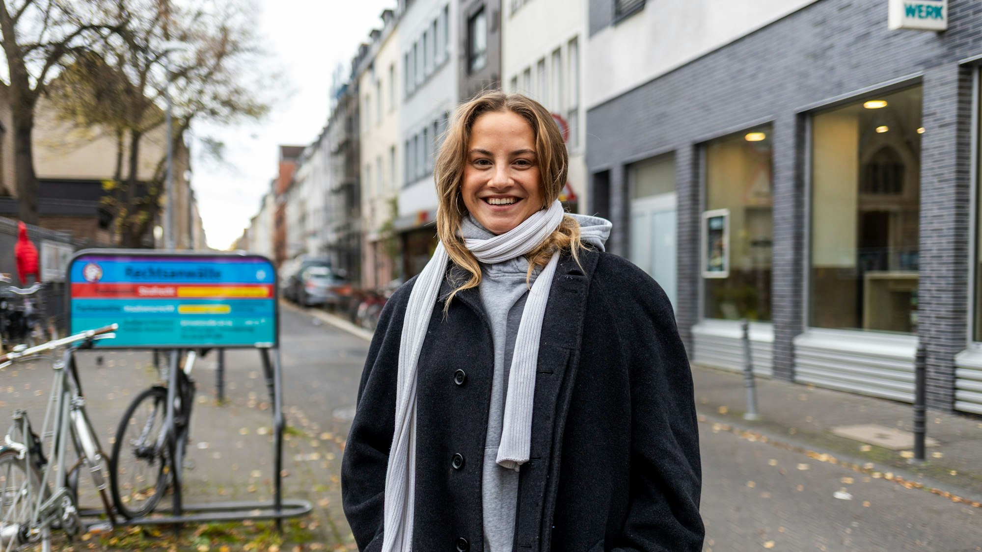 Eine junge Frau mit dickem schwarzen Mantel und weißem Schal steht auf dem Bürgersteig der Venloer Straße.