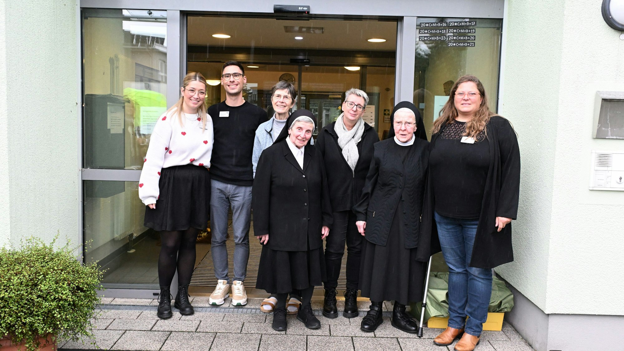 Zwei alte Ordensschwestern stehen mit anderen Menschen vor der Glastür eines Seniorenheims.