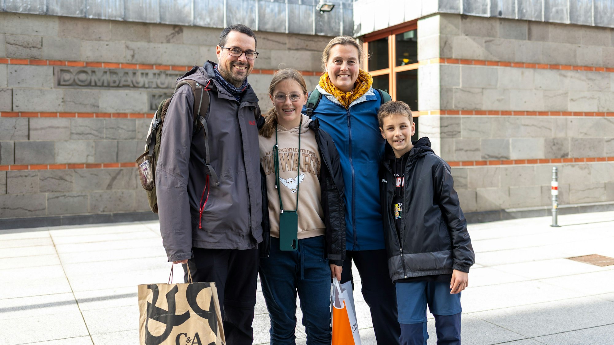 Vier Personen, ein erwachsener Mann, eine erwachsene Frau und zwei Kinder stehen vor der Rückseite des Kölner Doms. In den Händen halten sie Einkaufstüten.