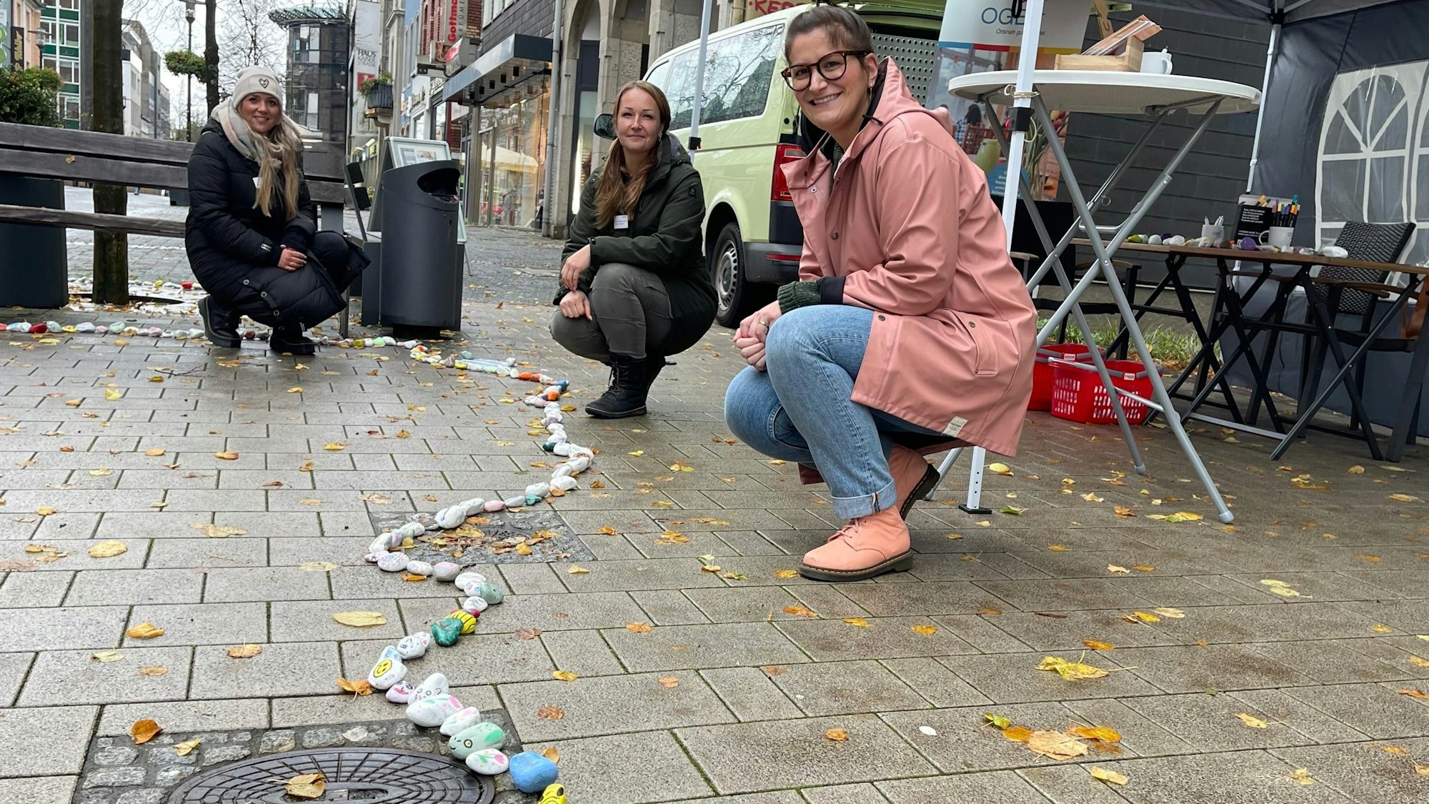 Drei Frauen Knien neben einer langen Schlange, die aus bemalten Steinen gelegt ist.
