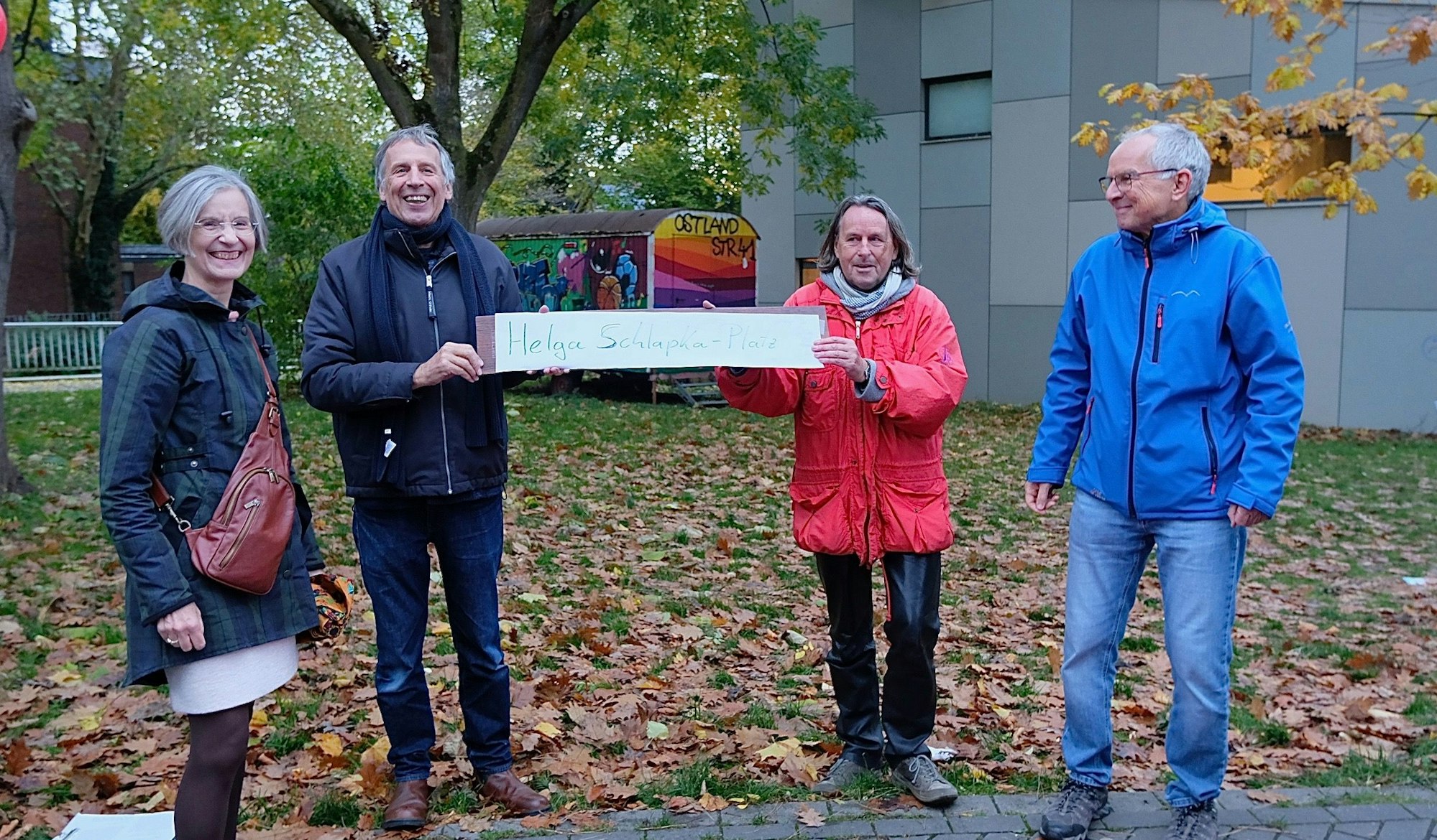 Eine Frau und drei Männer stehen an einer laubbedeckten Wiese. Zwei von ihnen halten ein handschriftlich gemaltes Schild mit der Aufschrift Helga-Schlapka-Platz.