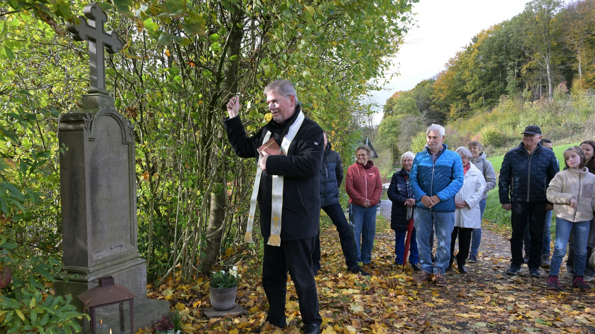 Ein Pfarrer segnet ein Wegekreuz. Dahinter stehen Menschen.