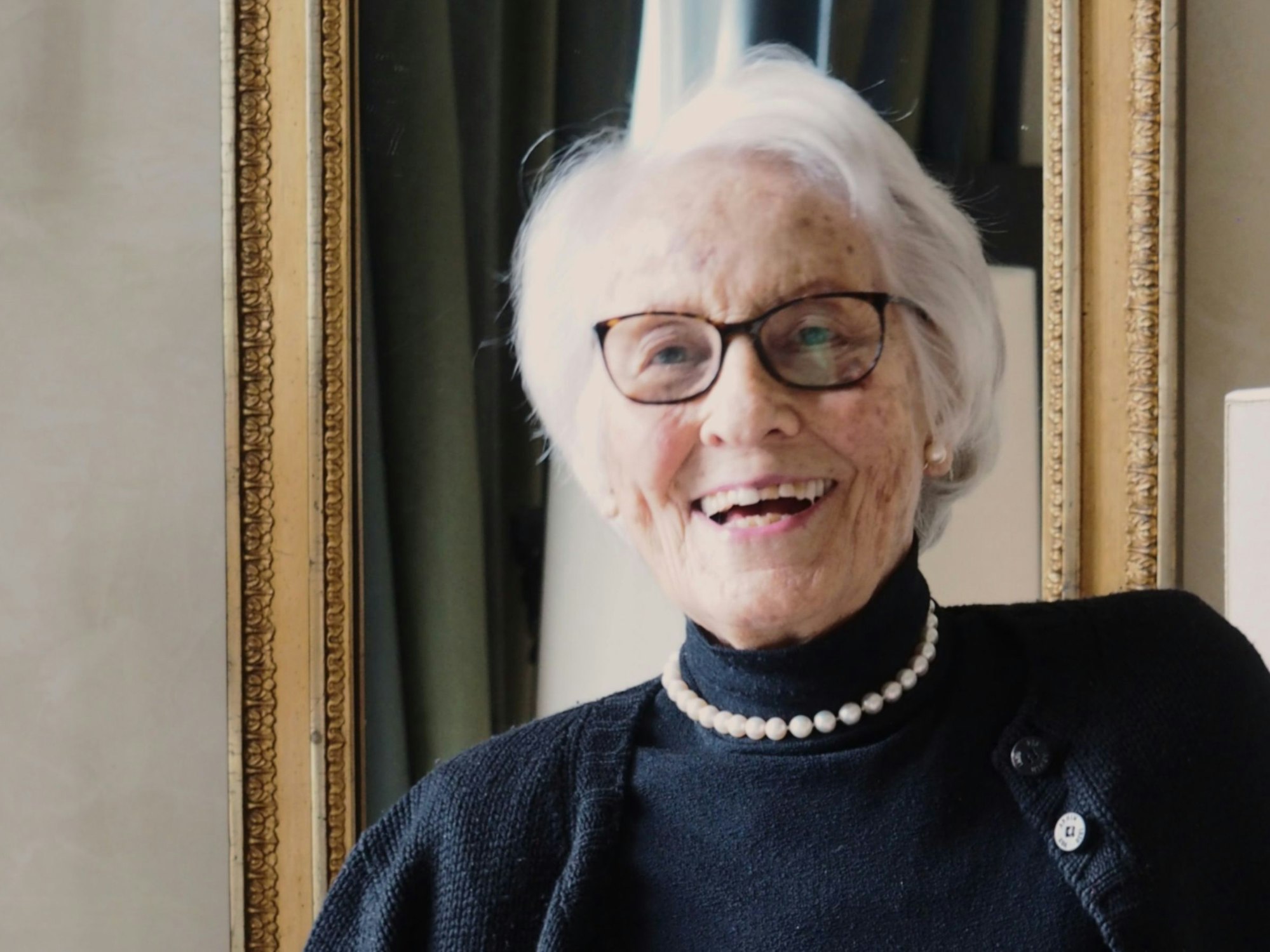 Eine ältere Dame mit kinnlangem Haar, Brille und Rollkragenpullover steht lachend in ihrem Wohnzimmer.