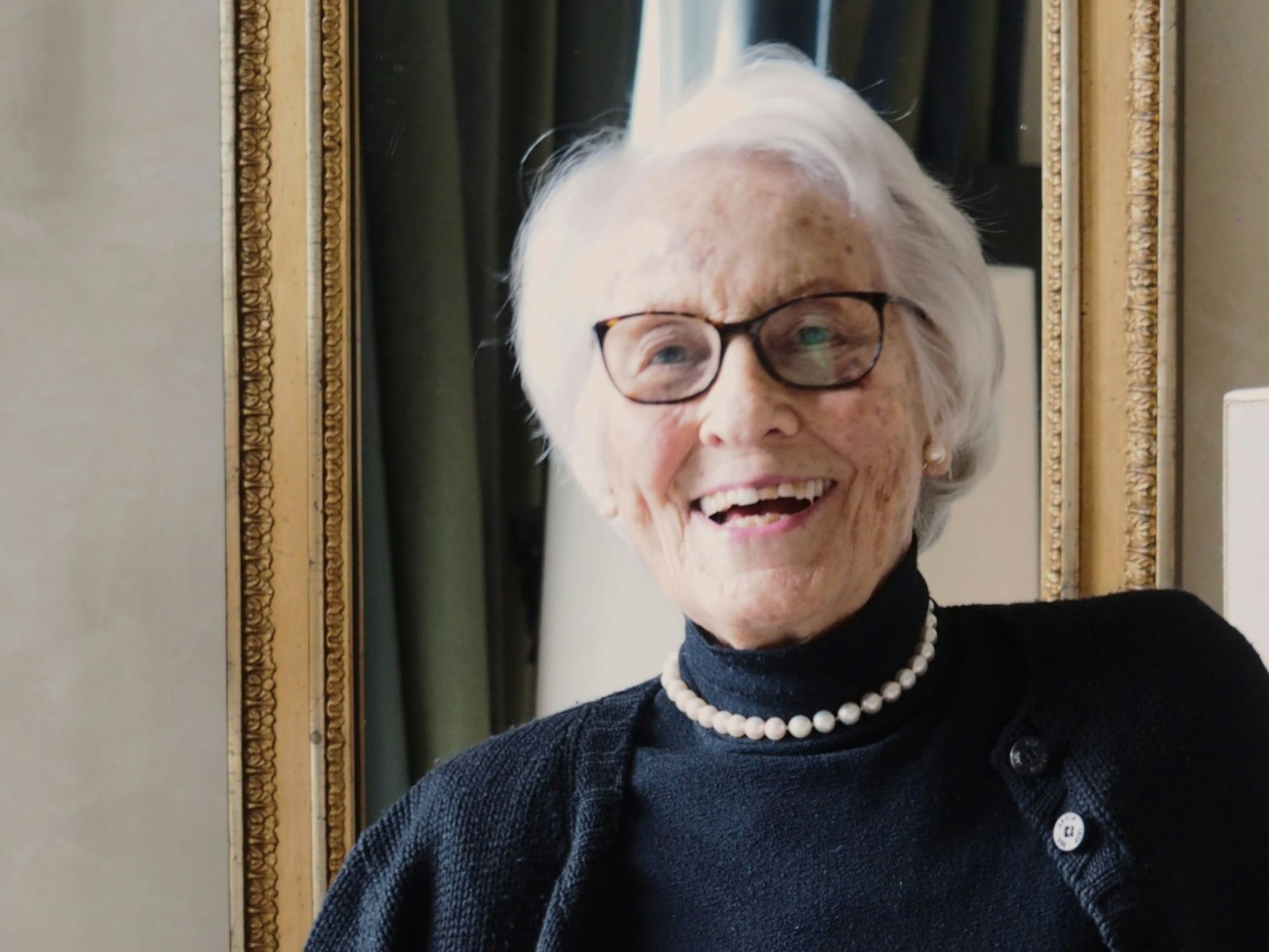 Eine ältere Dame mit kinnlangem Haar, Brille und Rollkragenpullover steht lachend in ihrem Wohnzimmer.