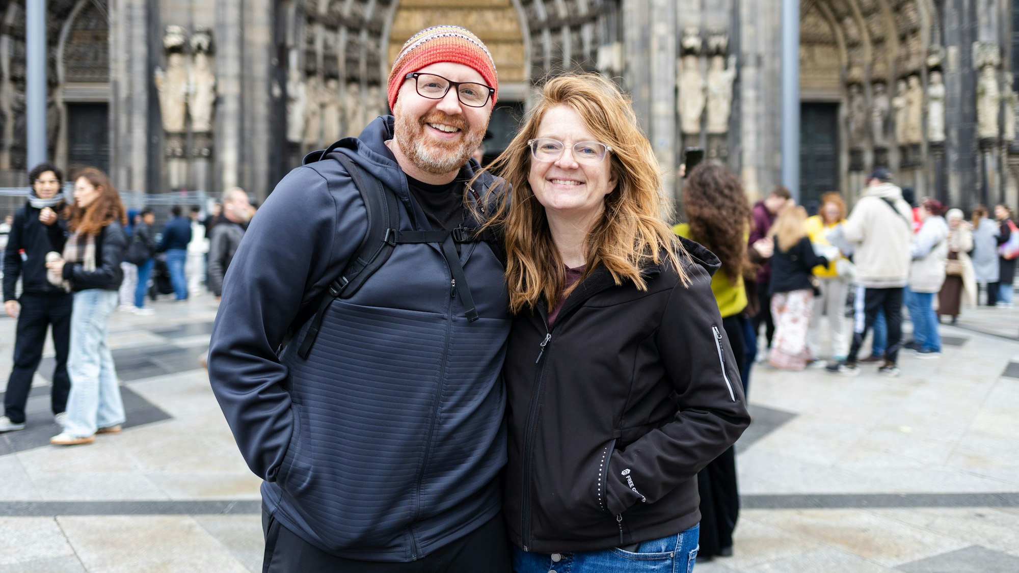 Ein Mann mir roter Mütze und eine Frau stehen Arm in Arm vor dem Kölner Dom.