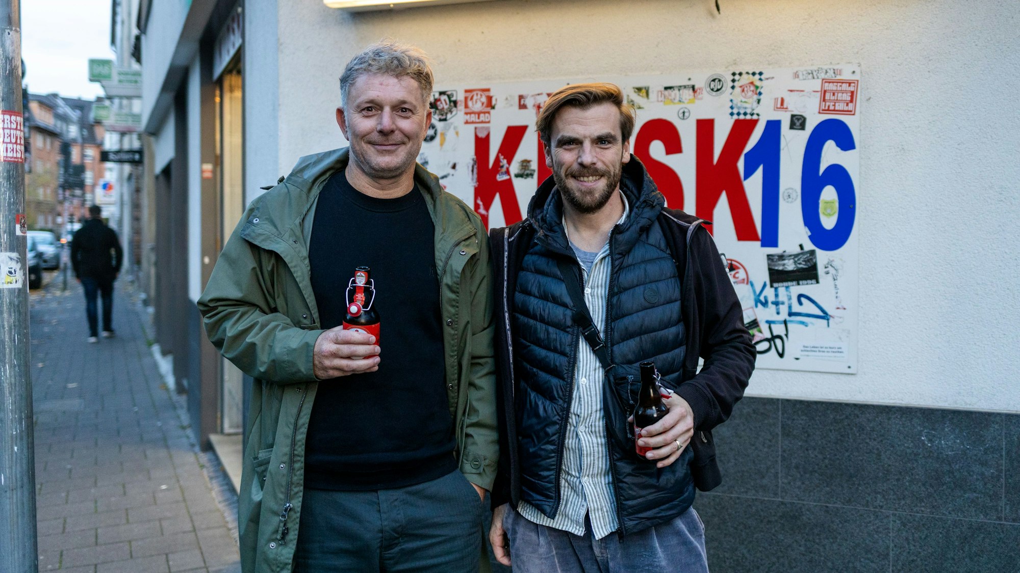 Zwei Männer mittleren Alters stehen mit Bier in der Hand vor einem Kiosk an der Deutzer Freiheit.