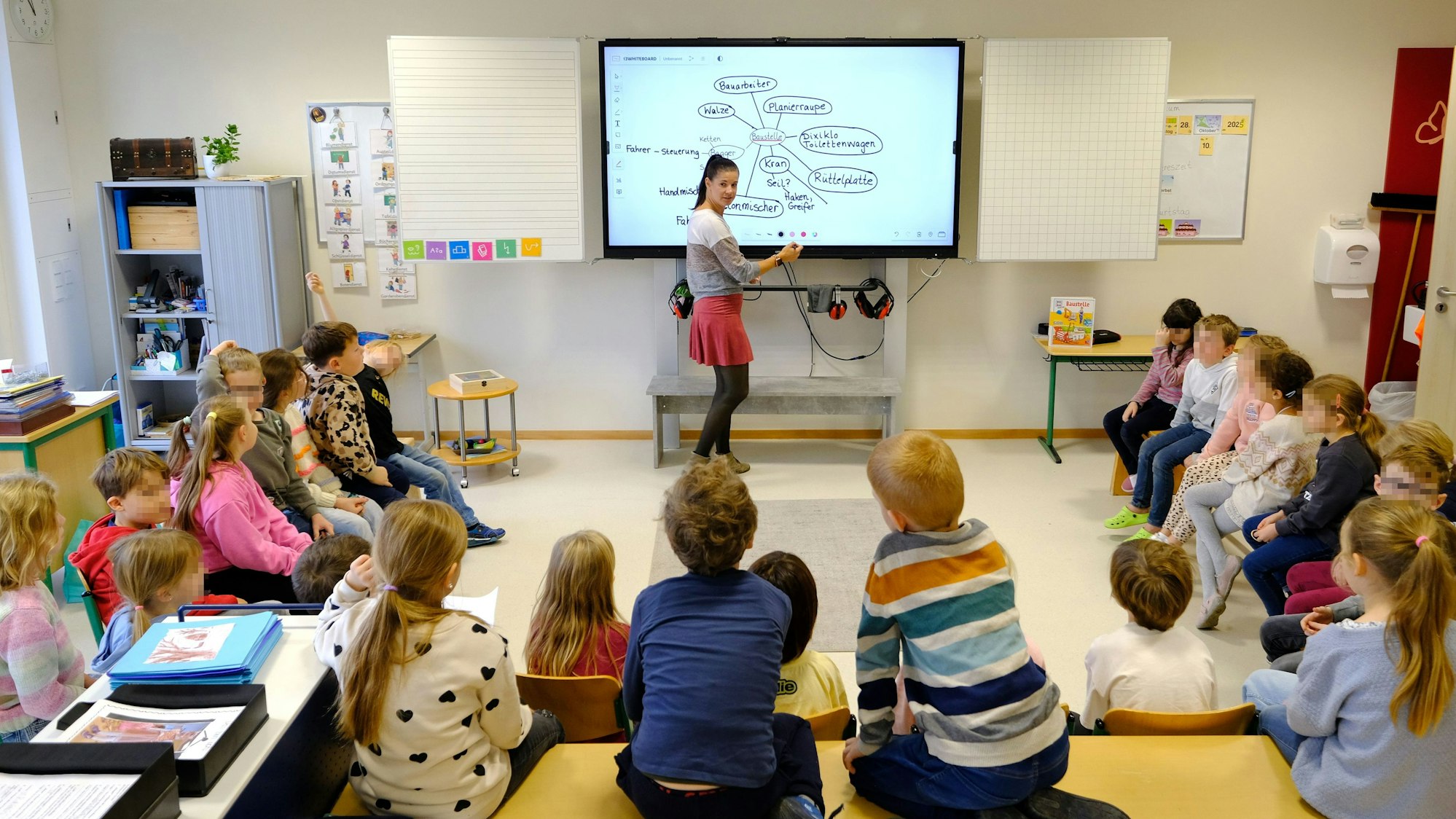 Blick in einen Klassenraum der Gemeinschaftsgrundschule Firmenich-Obergartzem. Die Lehrerin steht an der digitalen Tafel, die Schulkinder sitzen in einem Halbkreis davor.