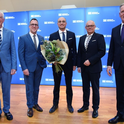 Fünf Herren im Anzug, der Mann in der Mitte hält einen Blumenstrauß.