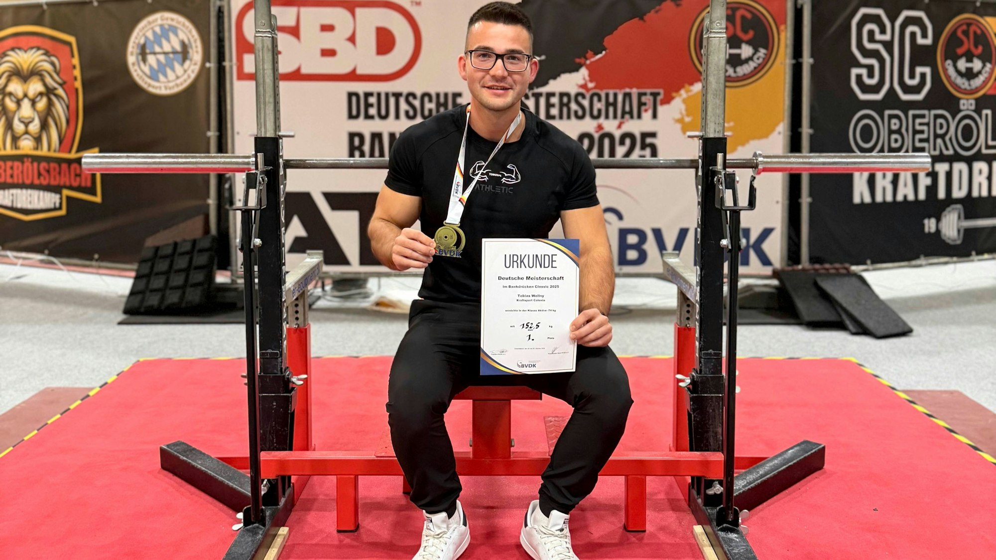 Ein Mann hält eine Urkunde in die Kamera, er sitzt auf einer Trainingsbank.