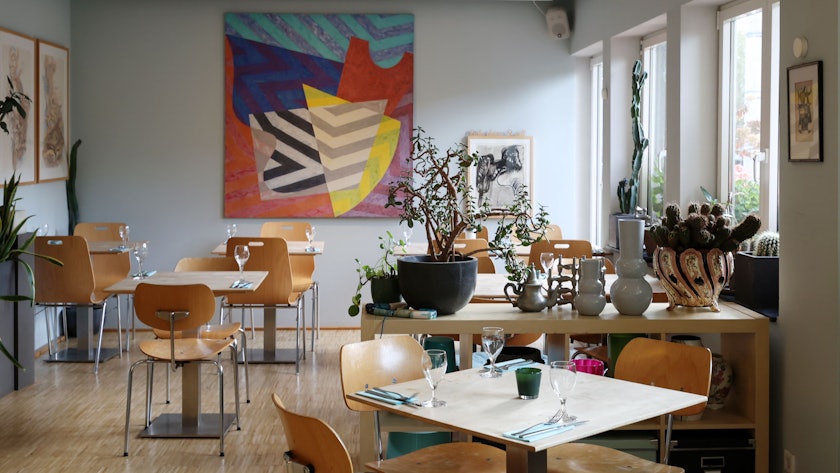 Innenraum des „Riphahn“: Holzstühle und Tische, eingedeckt mit Weingläsern, dekoriert mit Vasen, an der Wand hängt ein buntes Bild