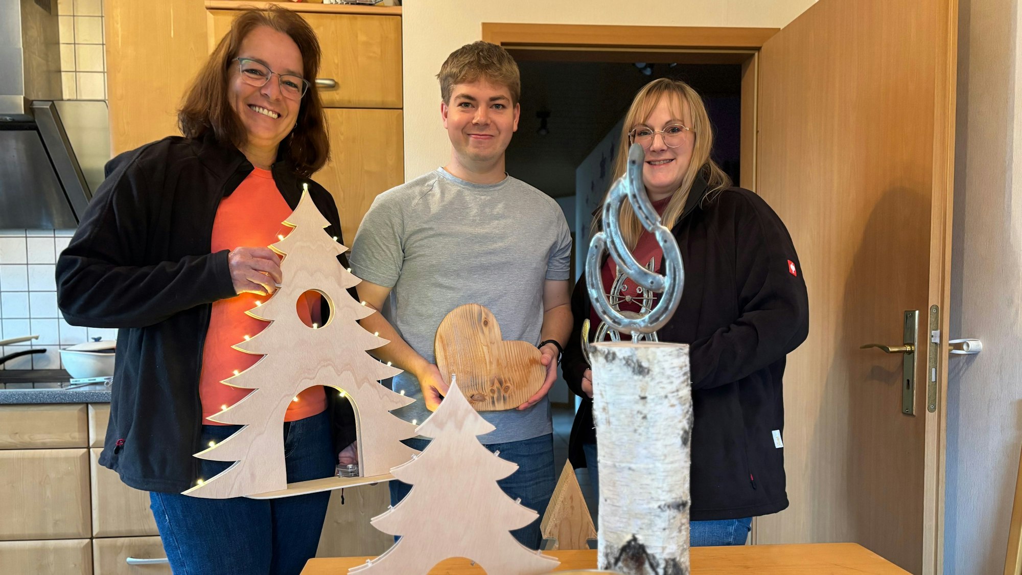 Michaela Lingscheid, Stefan Lingscheid und Ramona Bongartz stehen in einer Küche und halten hölzerne Dekorationsgegenstände in die Kamera.