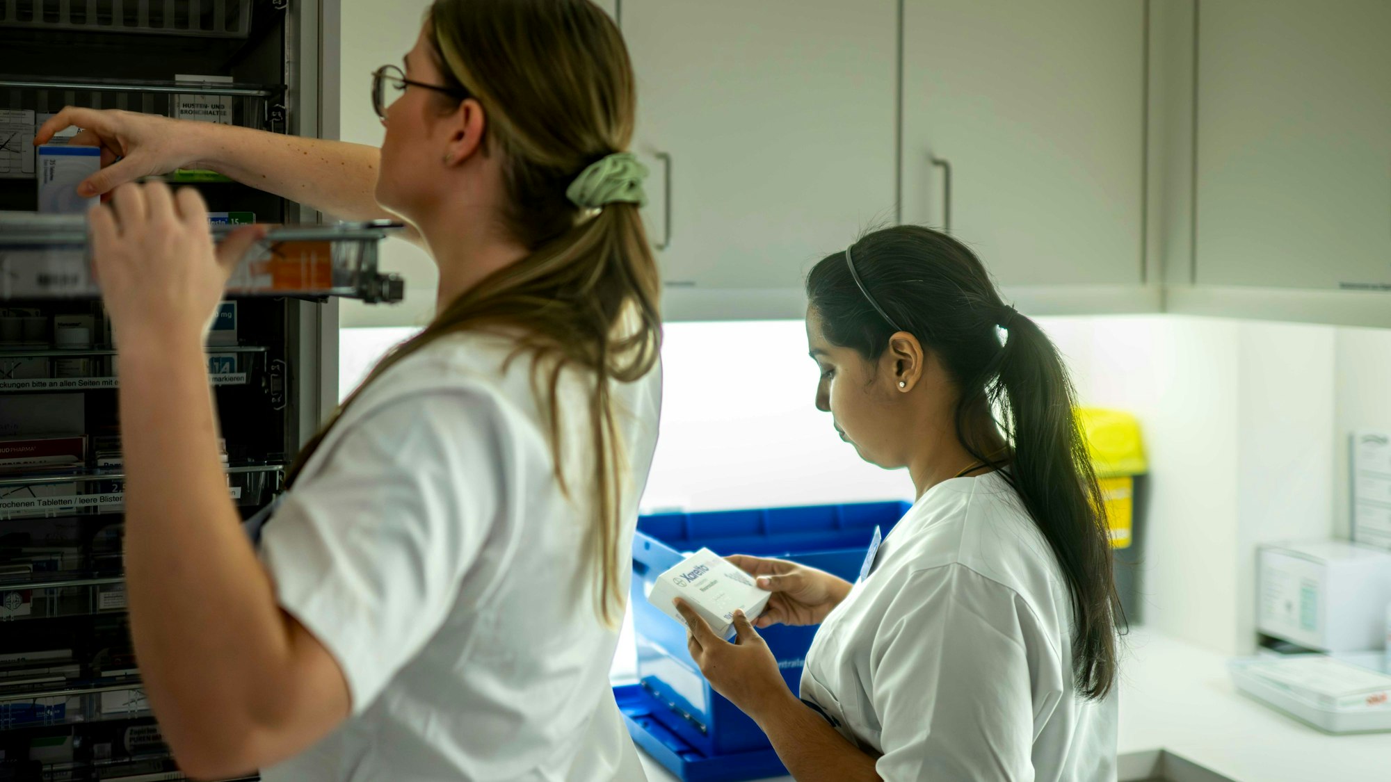 Eine Pflegekraft nimmt eine Medikamentenpackung aus dem Schrank, während ihre Kollegin auf eine andere Packung schaut.