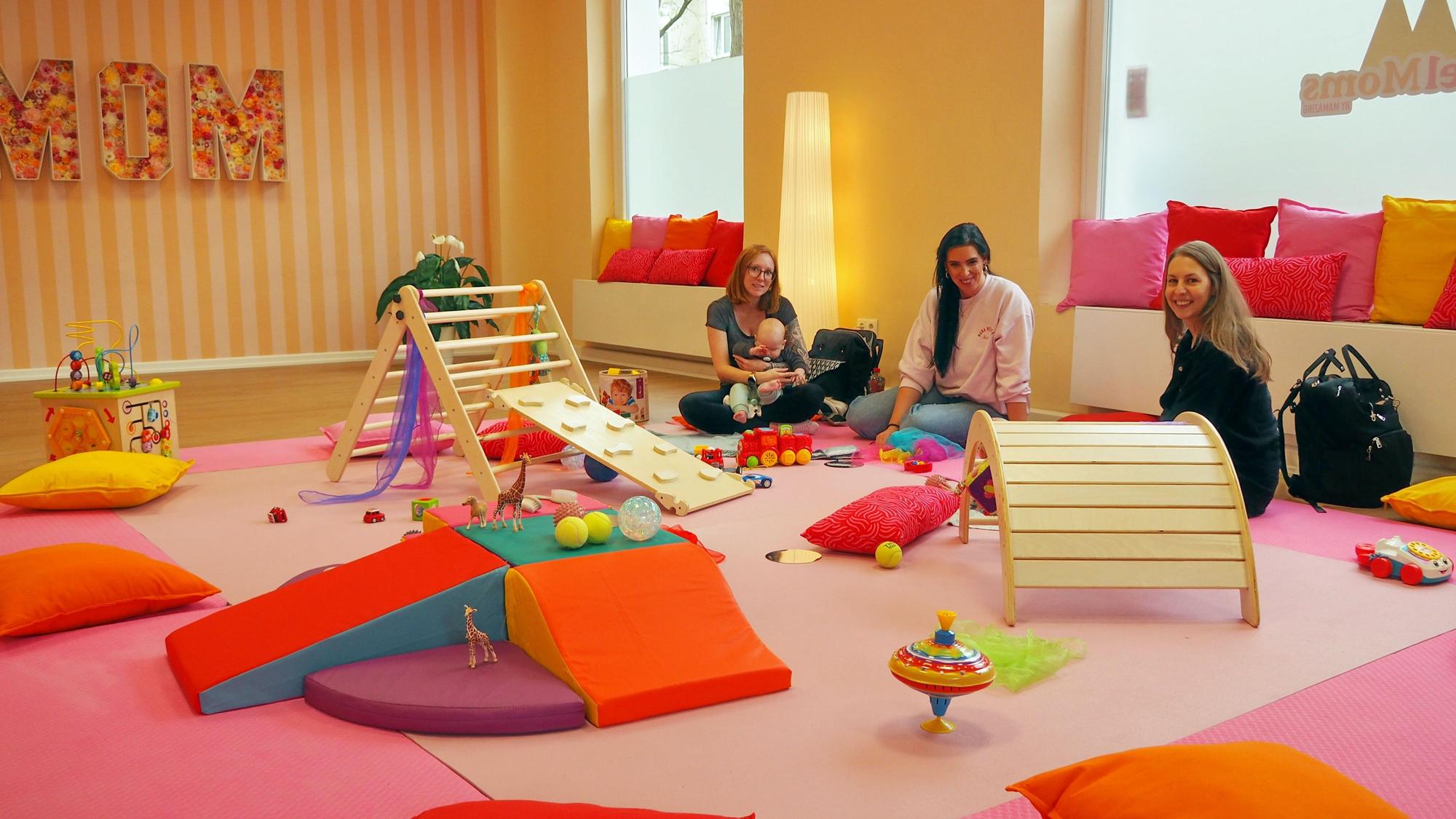 Drei Frauen sitzen in einem Raum voller bunter Matten und Spielgeräte für Kleinkinder.
