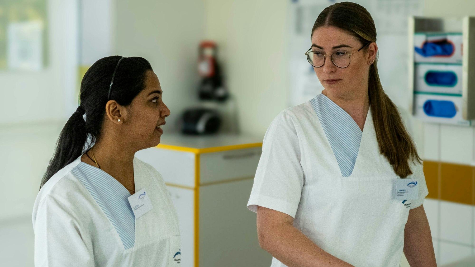 Die beiden Pflegekräfte des Marienhospitals tragen weiße Arbeitskleidung und unterhalten sich.