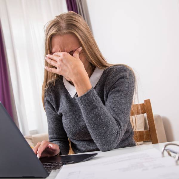 Eine Frau sitzt vor ihrem Laptop und hält die Hand vor Augen, weil sie Kopfschmerzen hat.
