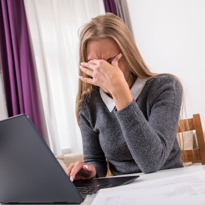 Eine Frau sitzt vor ihrem Laptop und hält die Hand vor Augen, weil sie Kopfschmerzen hat.