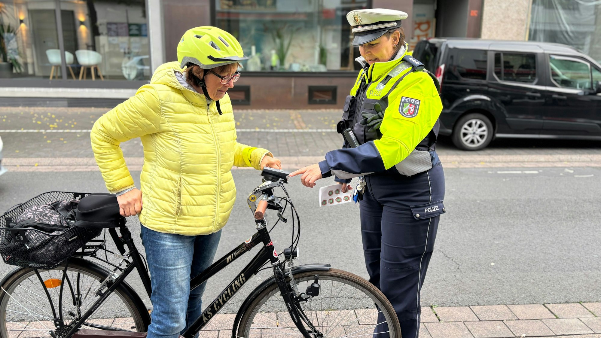 Ingrid Al-Hasan trägt eine auffällige helle Steppjacke und einen Fahrradhelm während sie neben ihrem Fahrrad steht. Anke Weber hat eine neonfarbene Warnweste und Polizeiuniform an und zeigt auf die Klingel des Fahrrads.