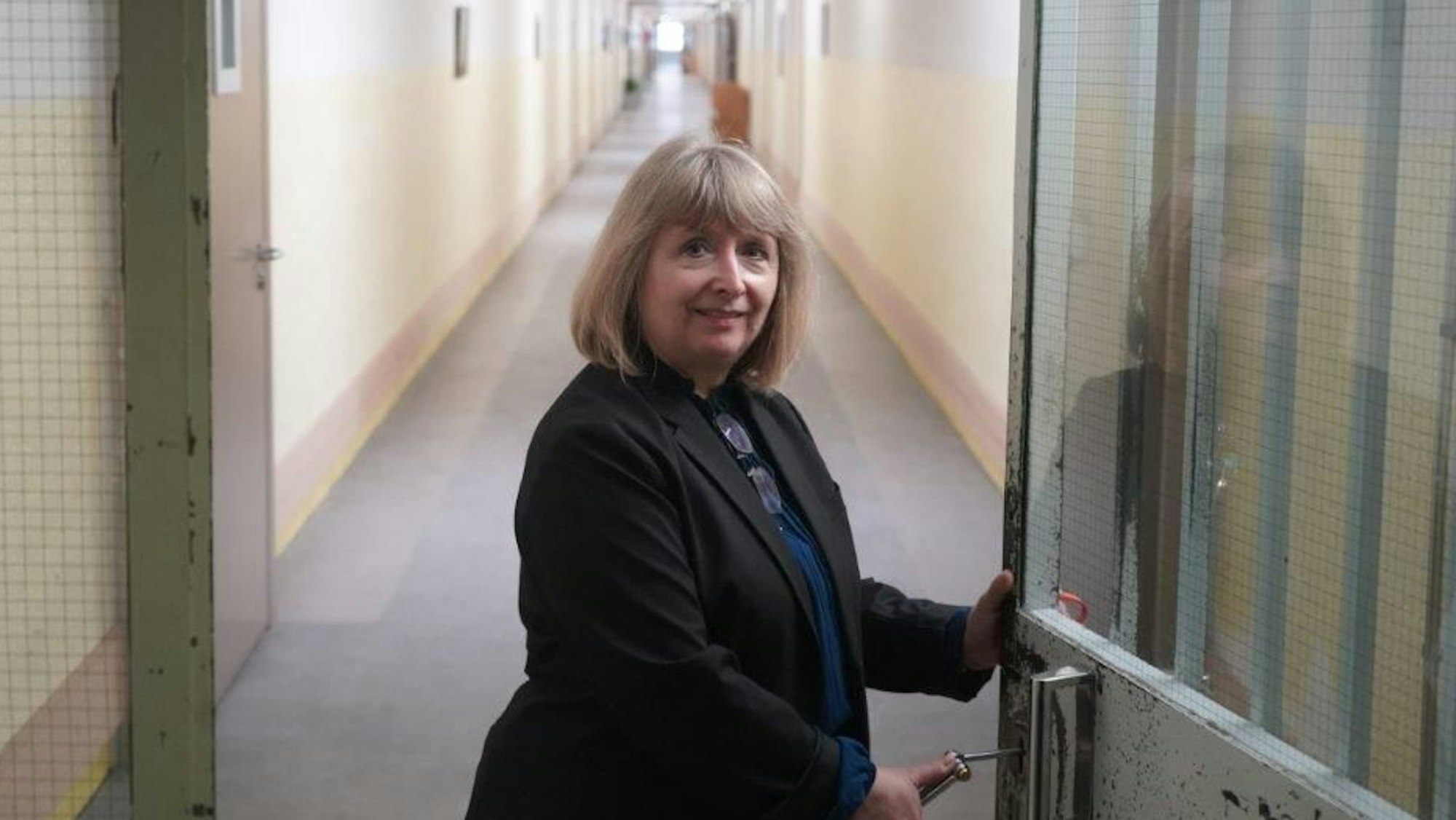 Eine Frau mit blondem schulterlangem Haar steht an einer Tür, hinter der ein langer Gang liegt.