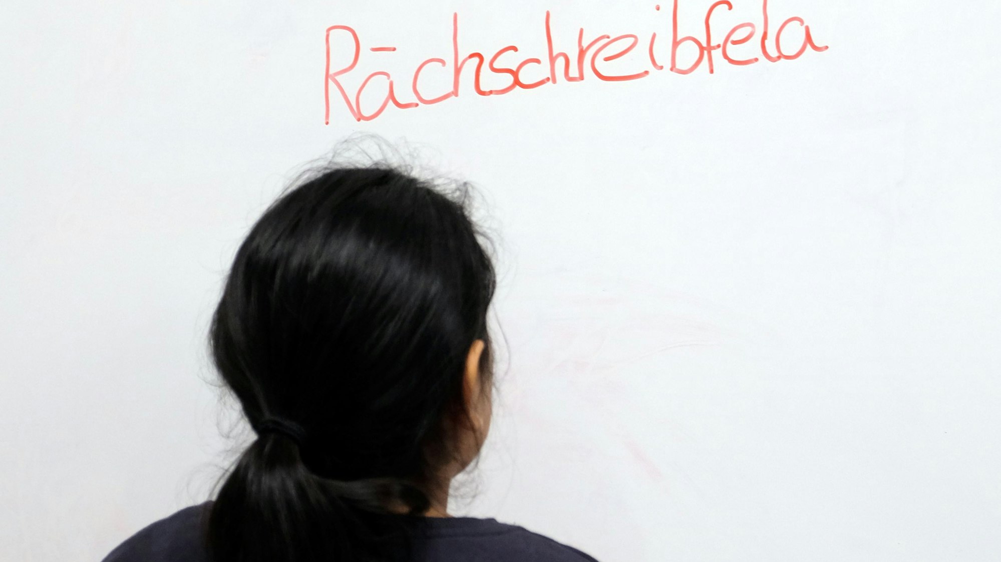 Ein Mädchen mit schwarzem Pferdeschwanz steht vor einer Tafel, auf der das Wort Rächschreibfela steht.