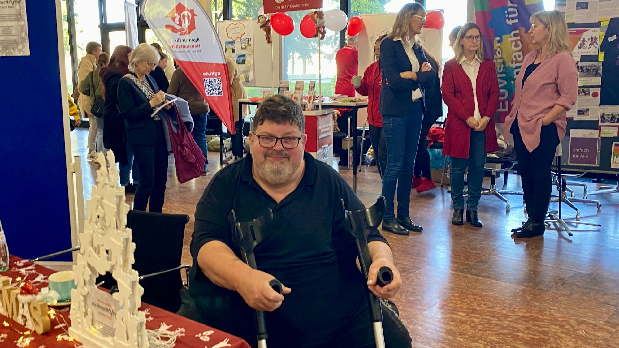 Klaus Berg sitzt an seinem Stand, in den Händen hält er Krücken. Im Hintergrund sind andere Stände. Menschen sind miteinander im Gespräch.