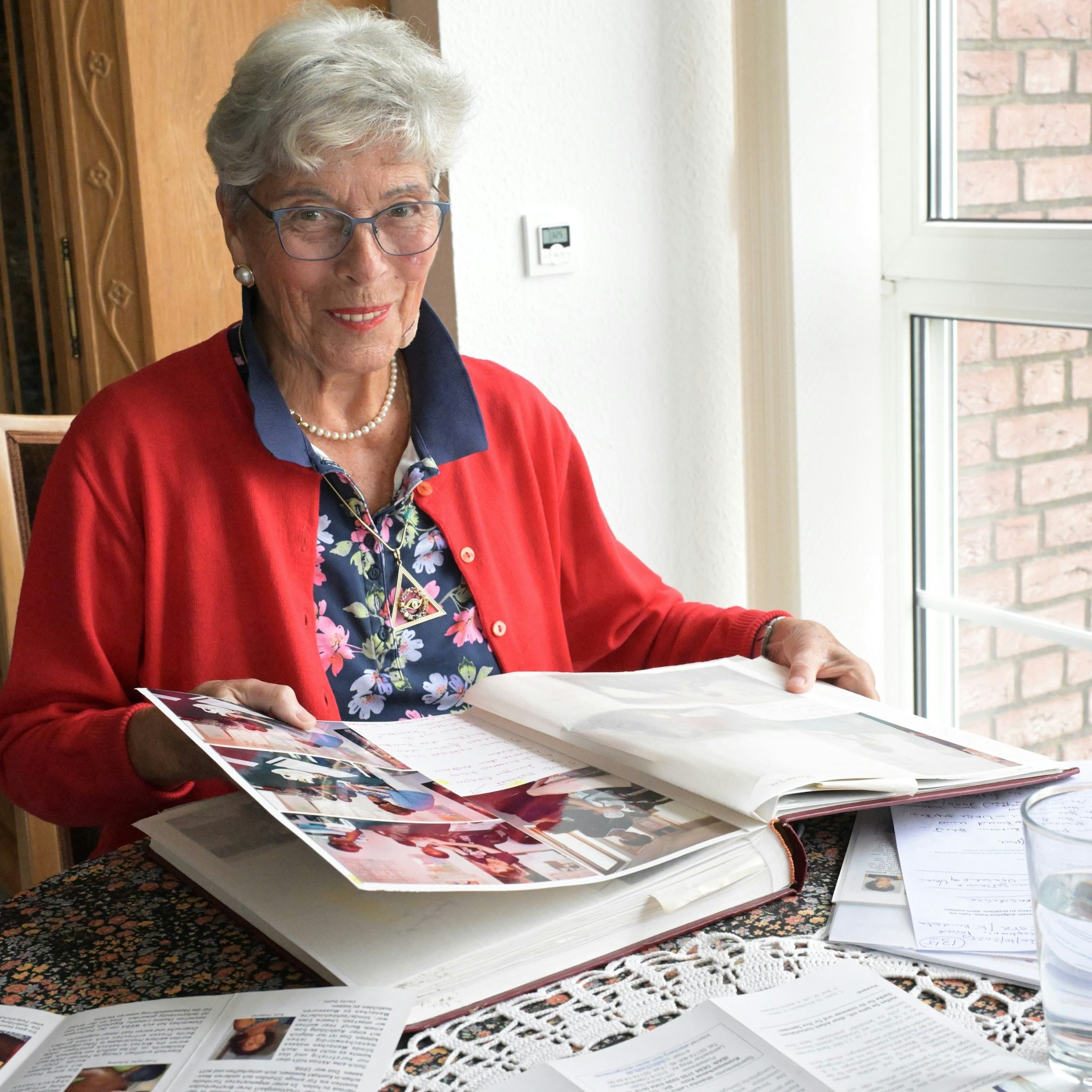 Eine ältere Frau in roter Strickjacke sitzt an einem Tisch und blättert in einem Fotoalbum