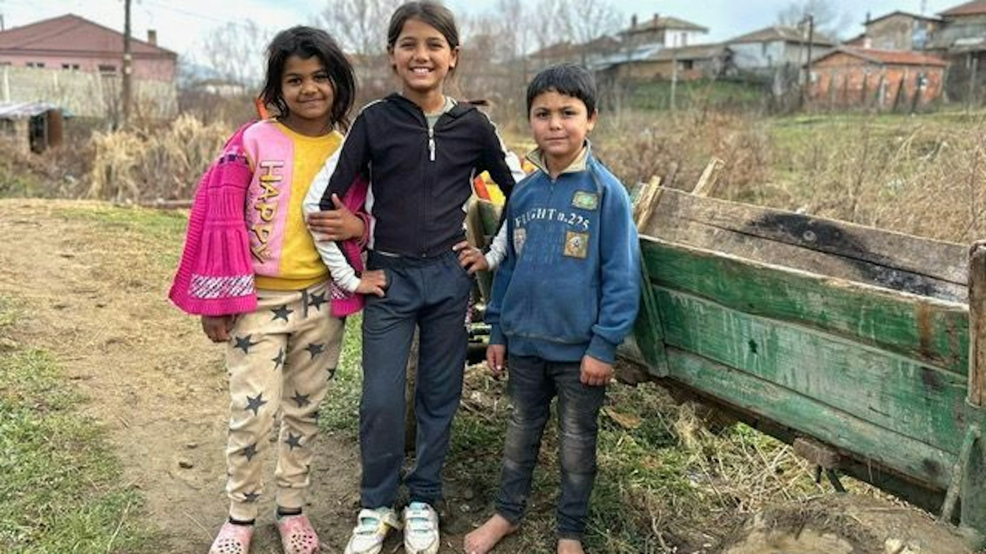 Zwei Mädchen und ein Junge stehen nebeneinander. Die beiden Mädchen lächeln. Der Junge hat keine Schuhe an.