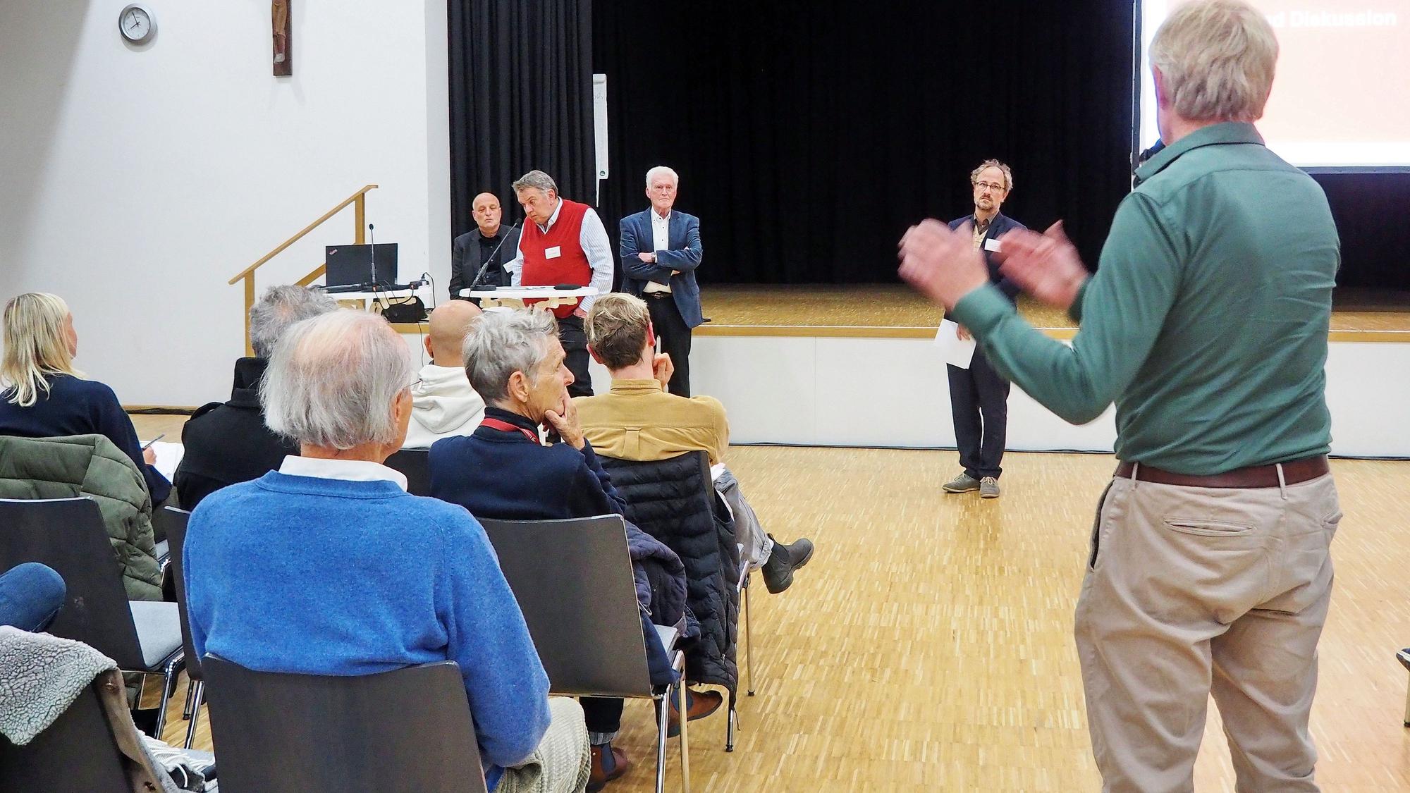 Menschen sitzen auf Stühlen in einer Schul-Aula.