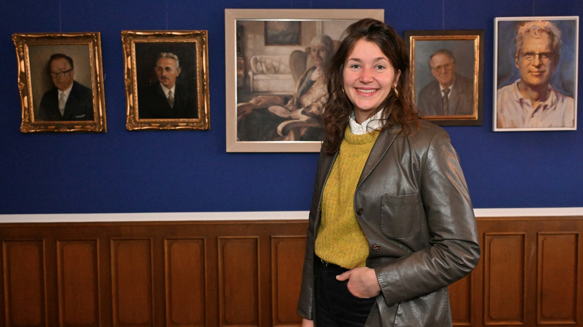 Eine junge Frau, Maja Supe-Dienes, steht lässig mit einer Hand in der Hosentasche vor einer blauen Wand mit Portraitgemälden von alten Menschen.