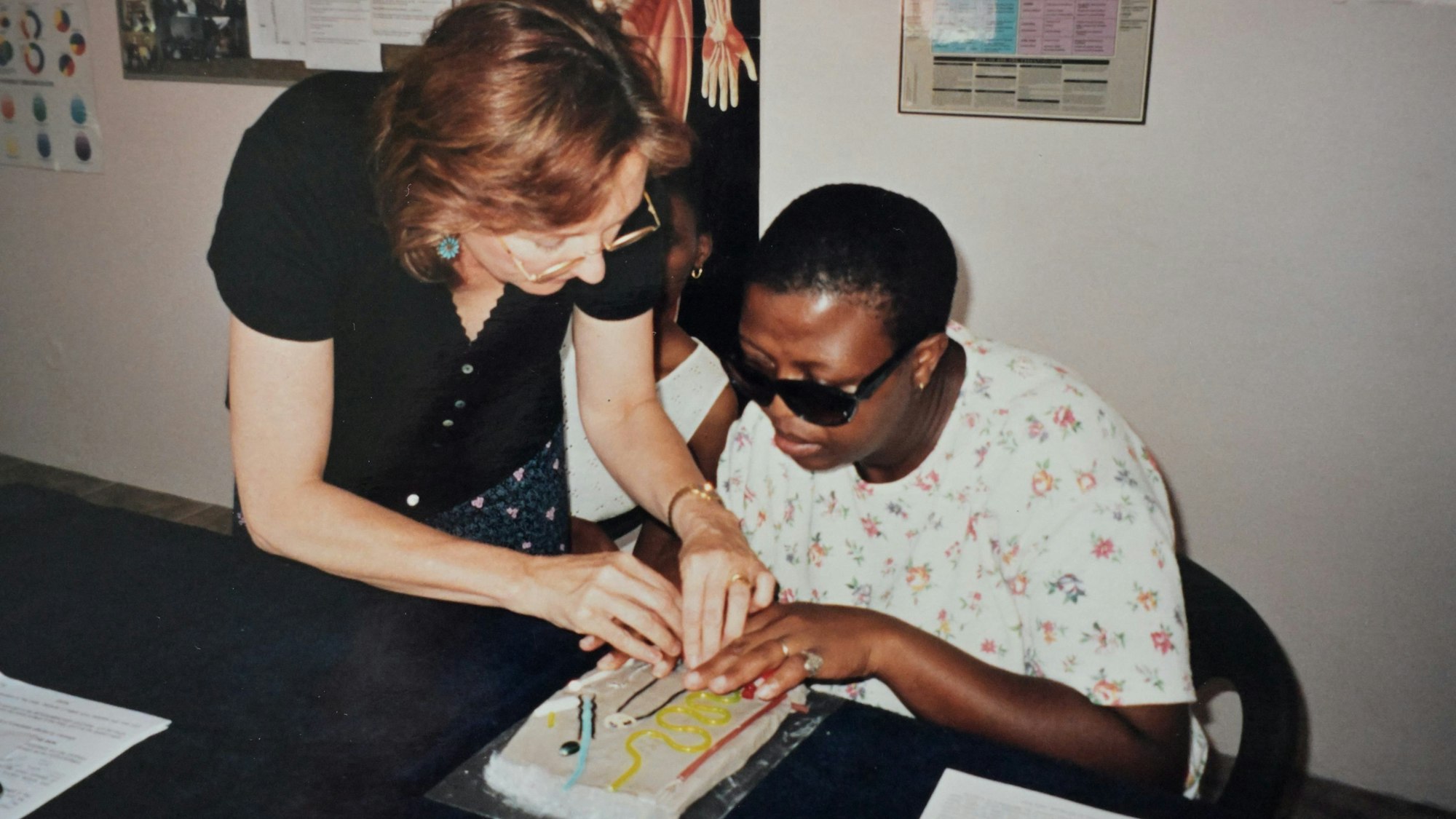 Eine blinde schwarze Frau sitzt an einem Tisch. Eine weiße Frau führt die Hände der Blinden über ein buntes Relief.